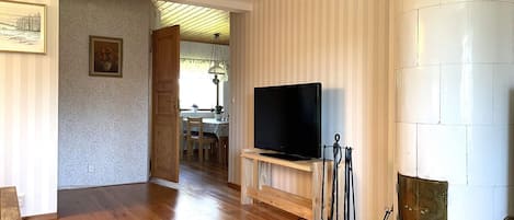 Uma sala de estar com piso de madeira, uma televisão sobre um móvel, uma lareira e uma área de jantar ao fundo.