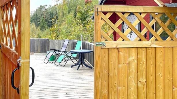 A wooden deck with a table and chairs, a red building in the background, and a wooden gate. - 5 Person Holiday Home in Ulrika (Ulrika)
