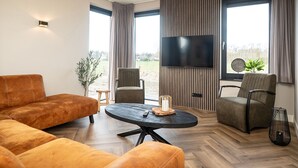 Una sala de estar moderna con sofá, sillas, una mesa redonda, televisor y ventanas amplias con cortinas.