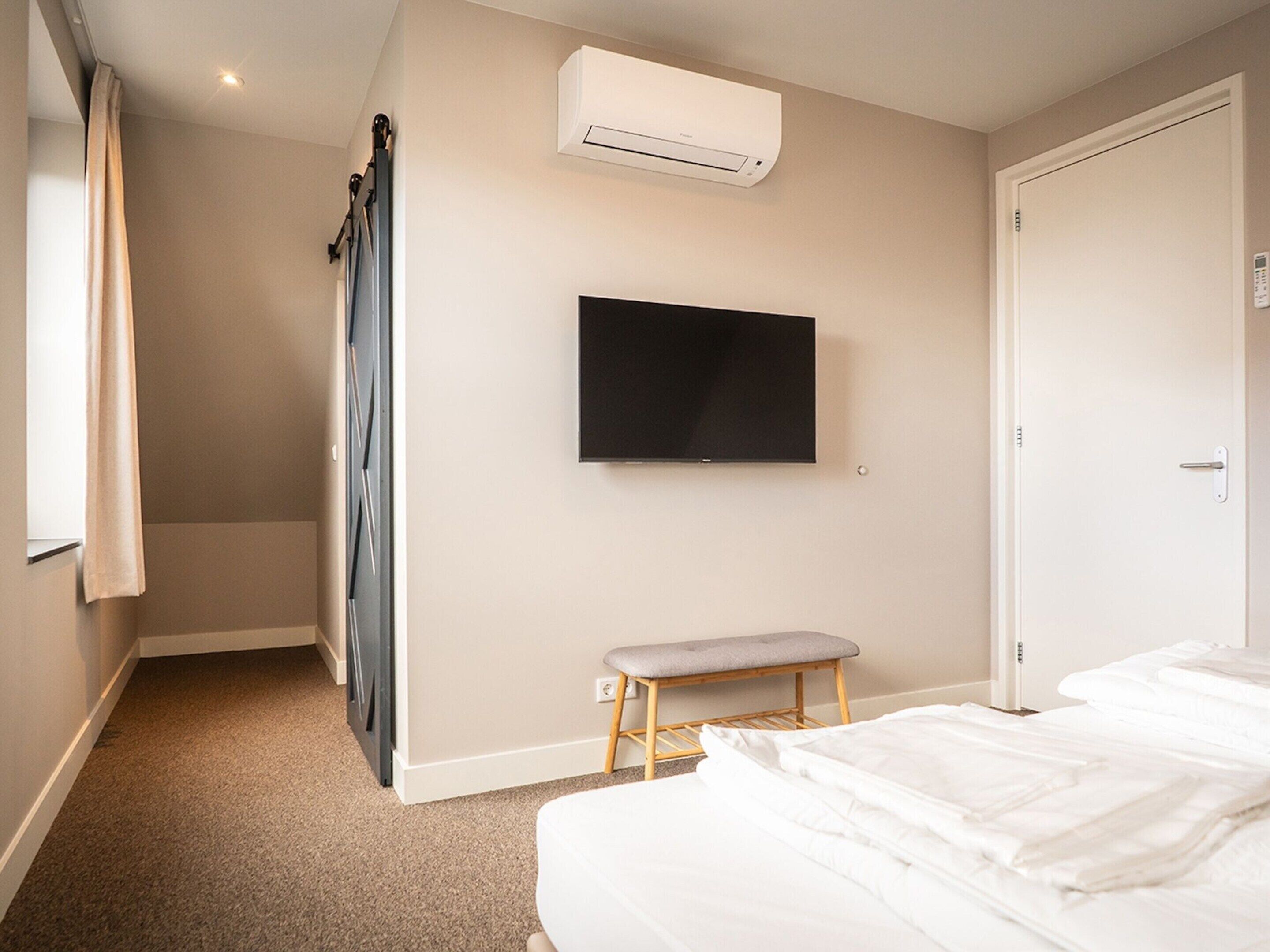 A bedroom with a bed, a TV mounted on the wall, an air conditioning unit, and a window with curtains.