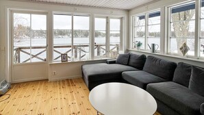 A room with a wooden floor, a gray sectional sofa, a white round table, and large windows overlooking a body of water. - 4 Star Holiday Home in Stensjon (Stensjon)