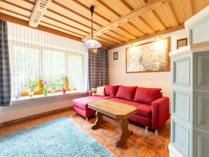 A cozy room with a red sofa, a wooden table, and a large window with curtains. - Apartment in Piesendorf Near Ski Slopes (Piesendorf)