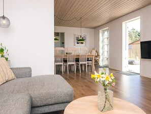 Ein modernes Wohnzimmer mit grauem Sofa, einem runden Holztisch mit Blumenvase und einem Essbereich mit Holzstühlen und einer grünen Pendelleuchte.