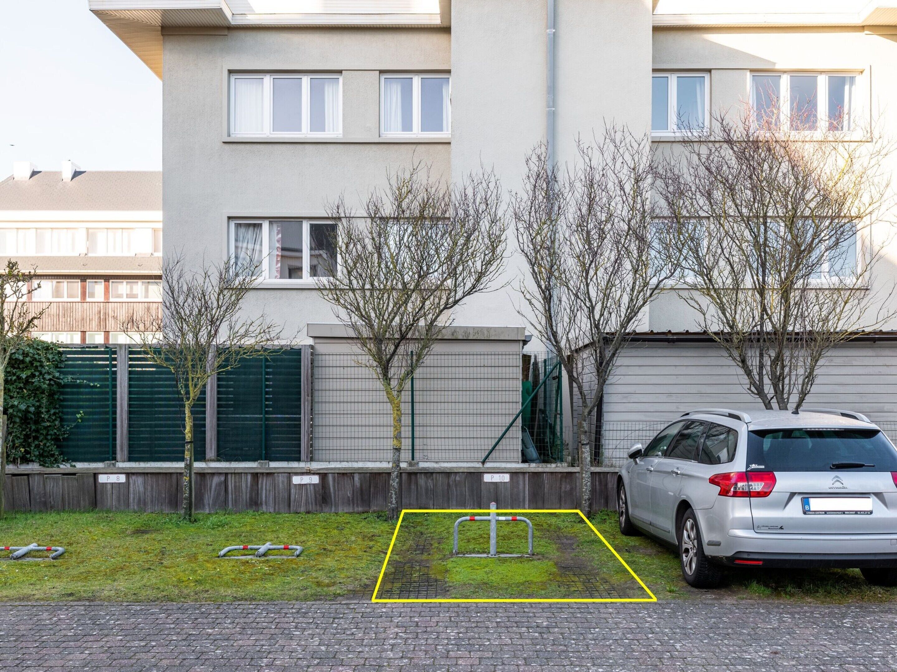 Ein Parkplatz mit einem Auto, einem markierten Stellplatz und einem Gebäude mit mehreren Fenstern im Hintergrund.