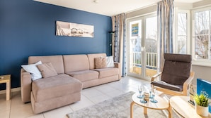 Un salon avec un canapé beige, un fauteuil marron, une petite table basse et une vue sur l’extérieur à travers une porte vitrée.
