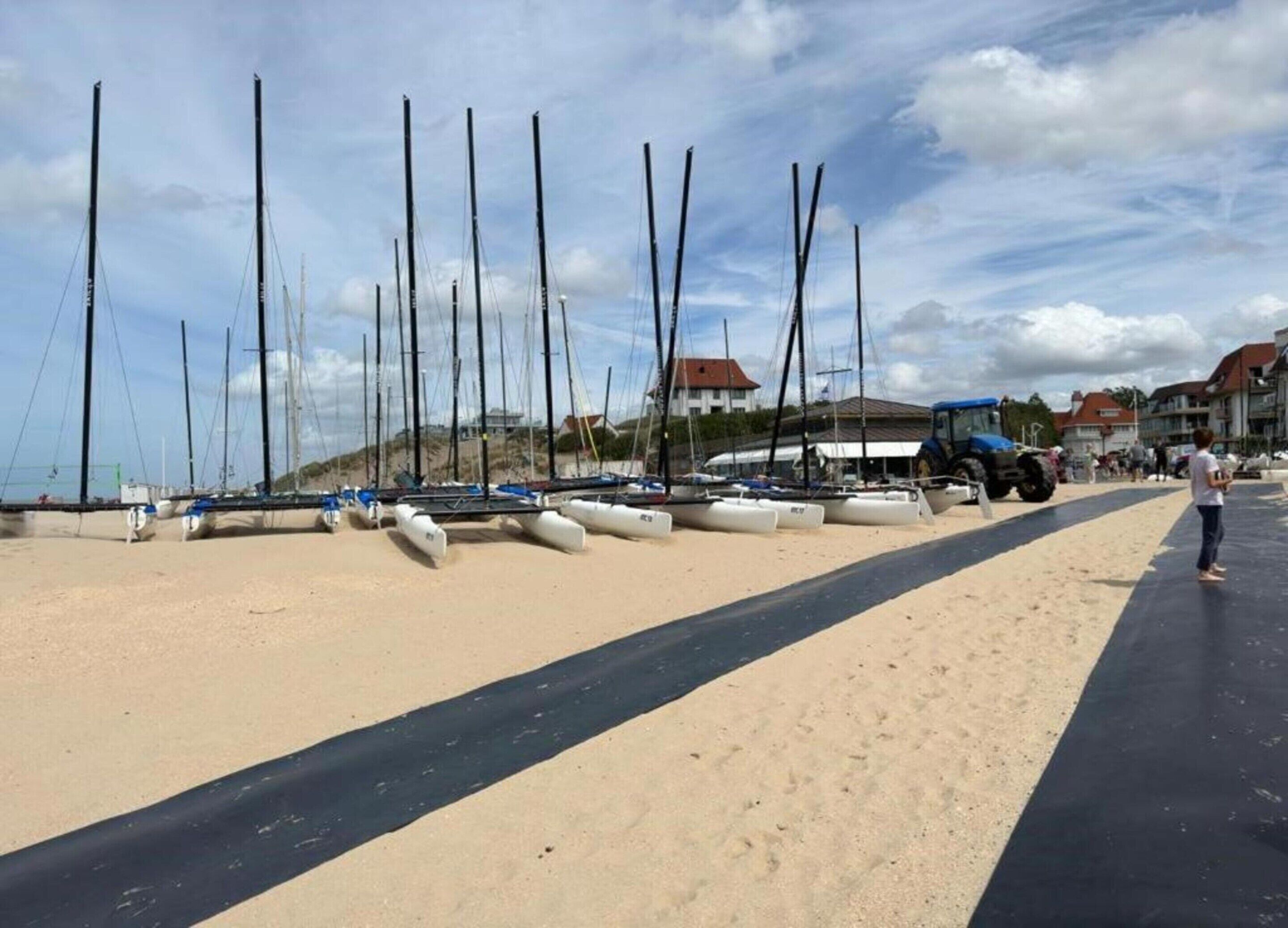 A sandy beach with multiple sailboats docked, a paved walkway, and buildings in the background.