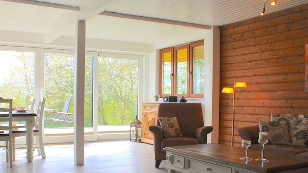 Ein geräumiges Wohnzimmer mit Holzboden, einer Couch, einem Sessel und einem Esstisch. Ein großes Fenster bietet Blick auf Bäume draußen.