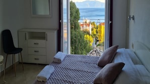 Un dormitorio con cama, escritorio y silla. Hay una ventana con vista al mar y edificios.