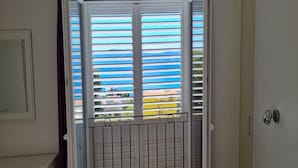 Una habitación con una puerta corrediza de vidrio, persianas horizontales, una pequeña reja metálica y vistas al exterior visibles a través de las persianas.