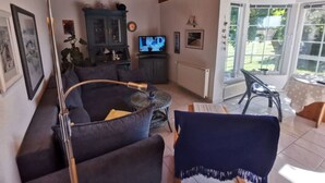 Un salon chaleureux avec un canapé bleu foncé, une petite table basse ronde et un fauteuil bleu. Un téléviseur est fixé au mur, et une fenêtre offre une vue sur l’extérieur.