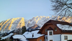 Ein Holzhaus mit geneigtem Dach und weißer Fassade, umgeben von verschneitem Boden und einem Bergmassiv im Hintergrund.