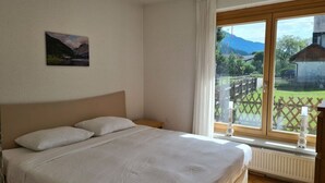 Ein Schlafzimmer mit einem Bett, einem Nachttisch und einem Fenster mit Blick auf ein Haus und Berge.