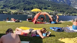 Ein Campingplatz am Seeufer mit Zelten, einem gelben Sonnenschirm und Menschen, die im Gras entspannen.