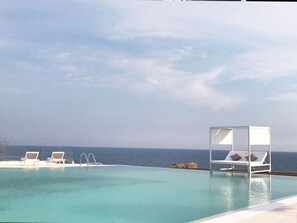 An infinity pool with clear blue water, white lounge chairs, and a canopy structure. - Apartment With Garden in Resort With Pool (Isola di Capo Rizzuto)
