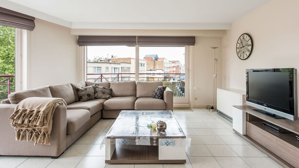 A modern living room with a sectional sofa, a coffee table, a flat-screen TV, and a large window with a view of buildings. - La Boheme 204 G1 Stylish Apartment in Koksijde (Koksijde)