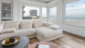 Un salon moderne doté d’un grand canapé d’angle, d’une table basse ronde et offrant une vue sur la mer et un bâtiment remarquable, le tout visible à travers de grandes fenêtres.