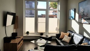 Una sala de estar con un sofá de cuero negro, una mesa de centro de vidrio, un mueble bajo para televisión de madera y un ventanal con persianas.