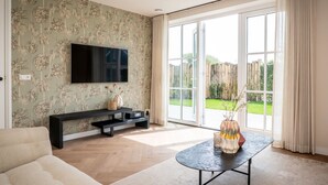A living room with a large TV, a sofa, a coffee table, and a view of a garden through a sliding glass door. - Villa on Texel Near Beach and Forest (De Koog)