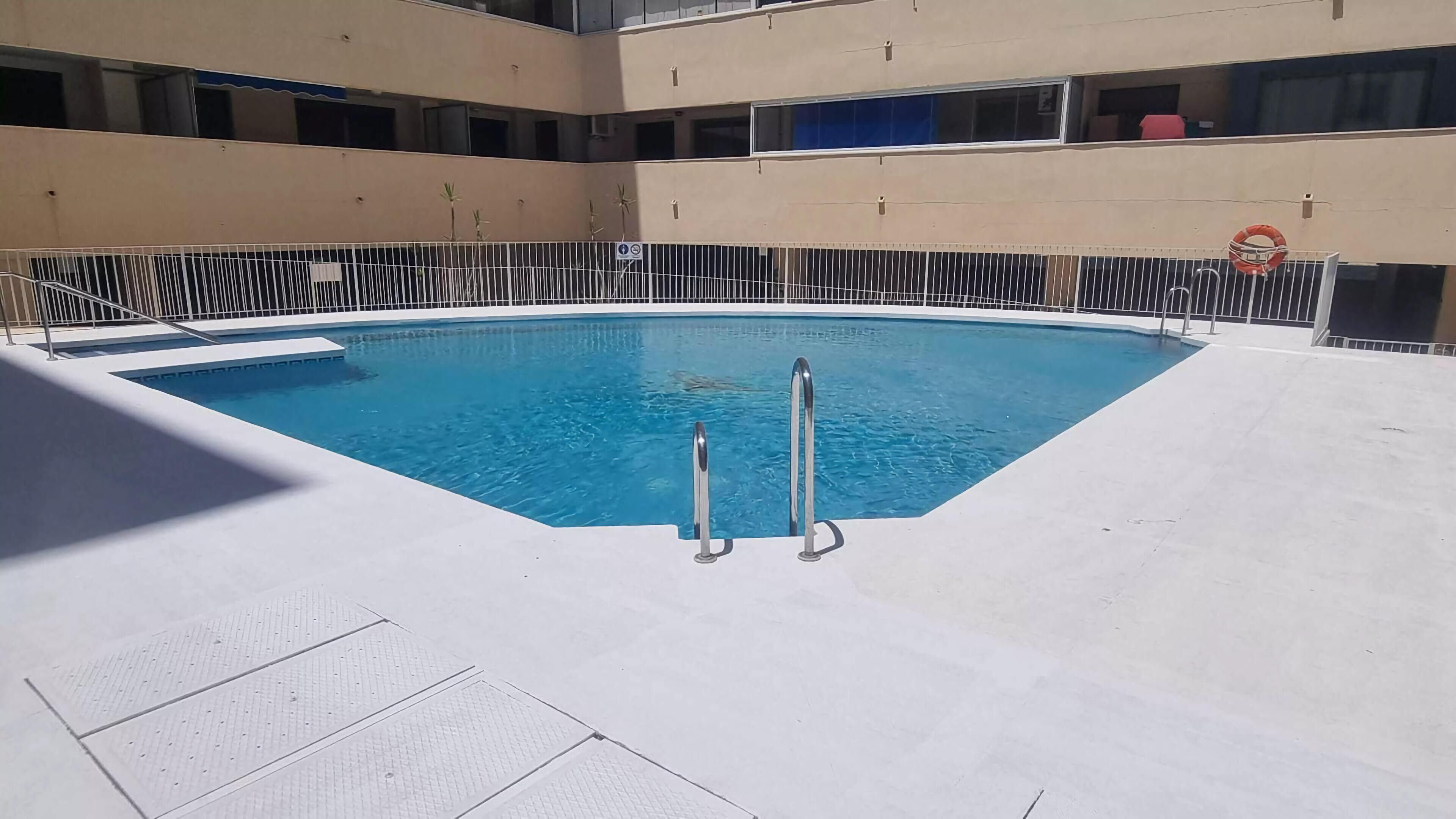 Una piscina con dos escaleras, rodeada por un deck de baldosas blancas y una cerca de metal.
