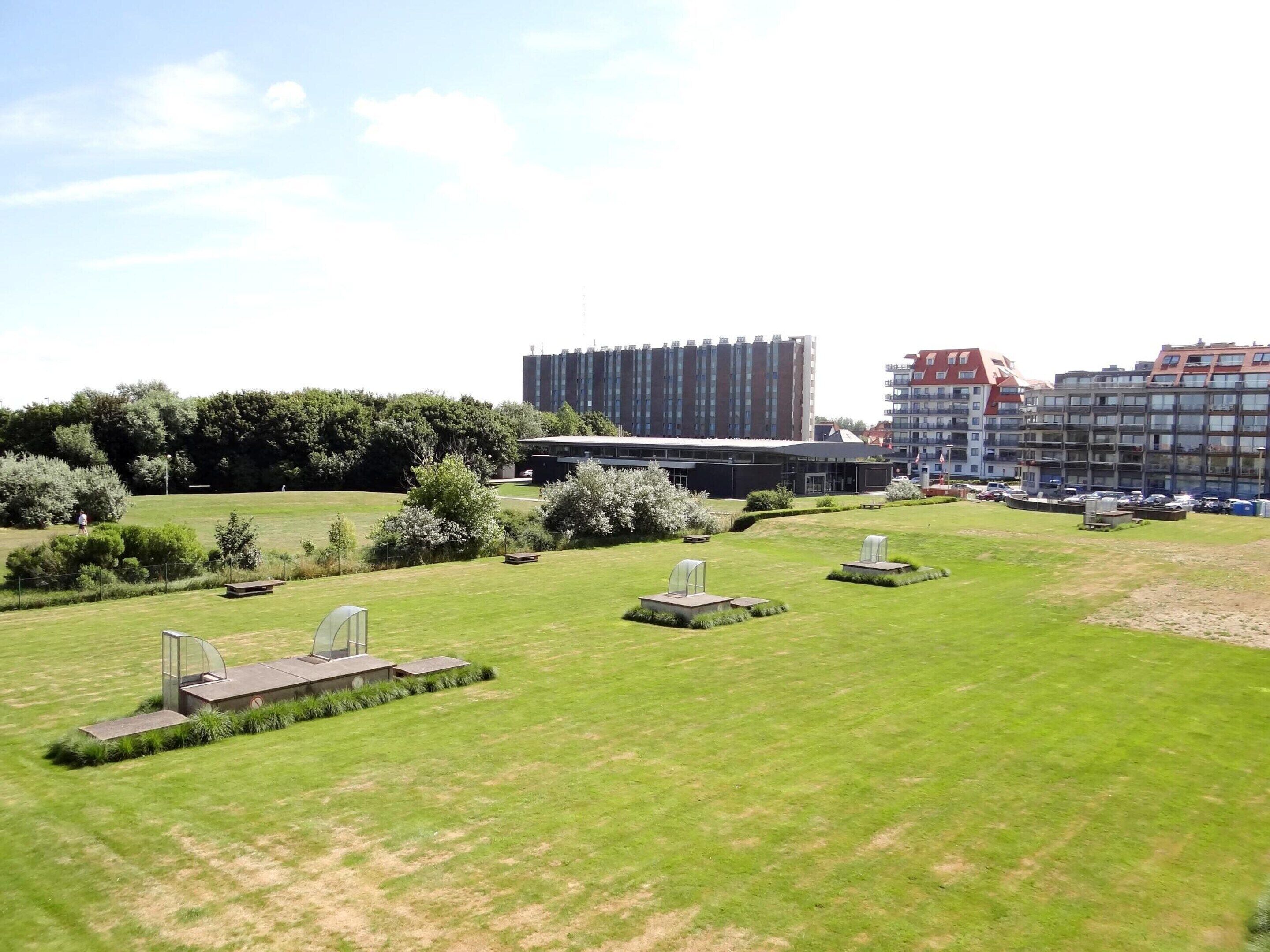 A grassy area with several headstones, a modern building, and residential buildings in the background.