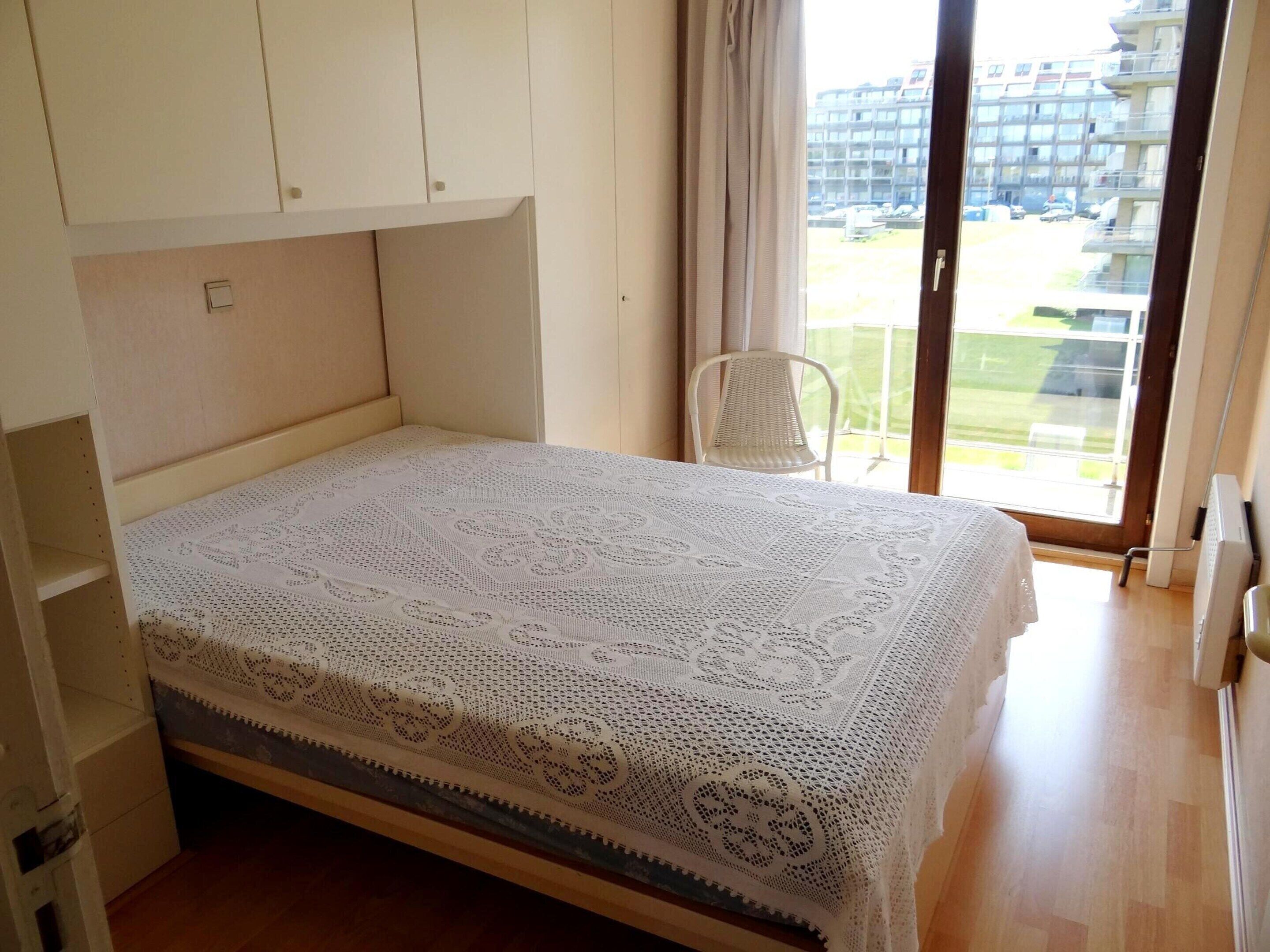 A bedroom with a bed covered in a white lace bedspread, a white chair, and a view of a building through a window.