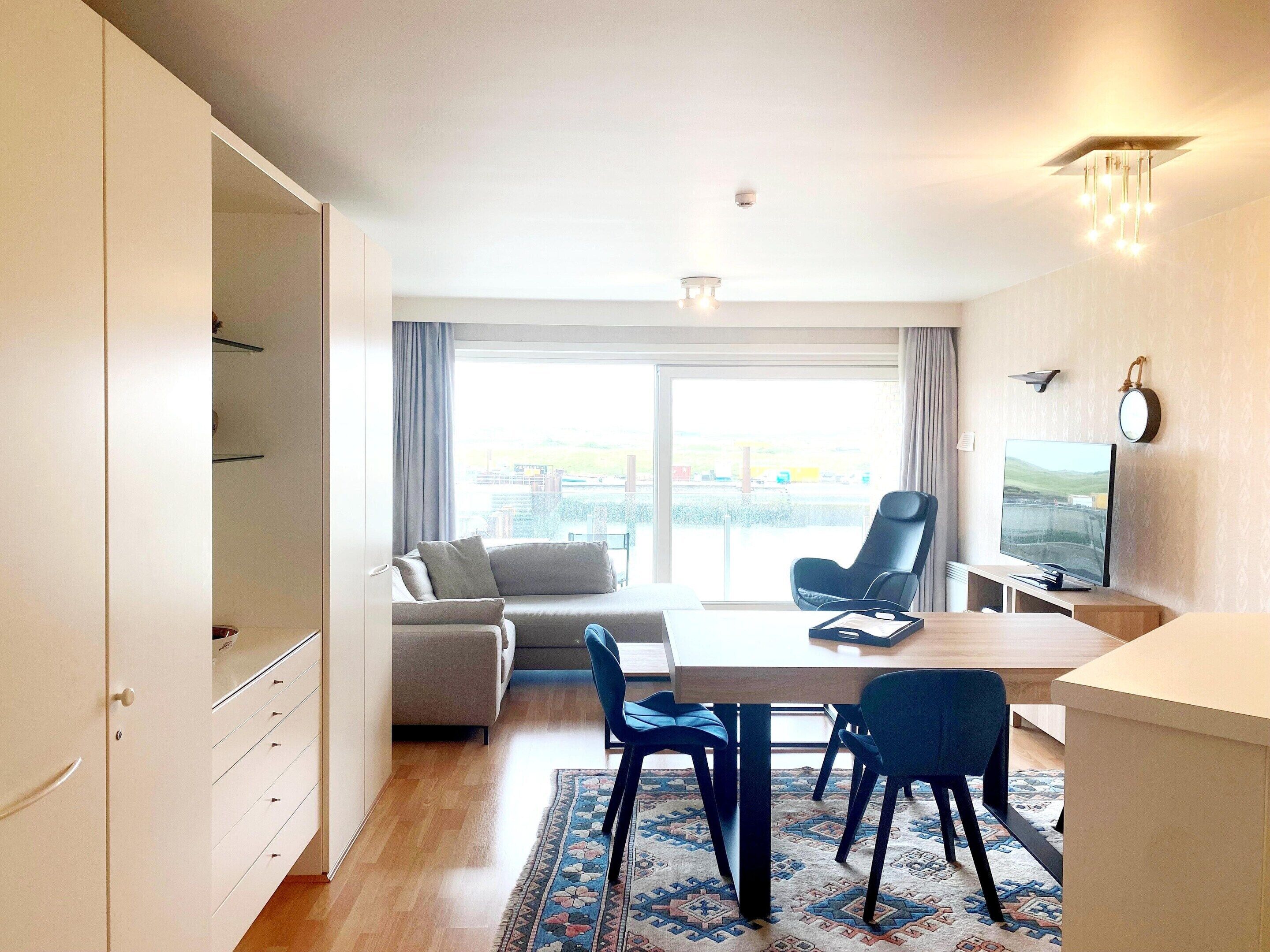 Una sala de estar moderna con un sofá, una mesa de comedor con sillas, un televisor y un gran ventanal con cortinas.