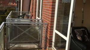 Een balkon met een houten terras, een glazen balustrade en een schuifpui die uitkomt op een kamer met een zwarte bank en gestreepte kussens.