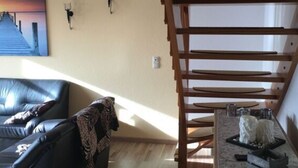 Een woonkamer met een zwarte leren bank, een houten trap, een schilderij van een zonsondergang en twee wandlampen.