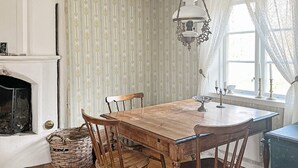 Une salle à manger avec une table en bois, quatre chaises, un lustre, une cheminée et une fenêtre avec des rideaux.