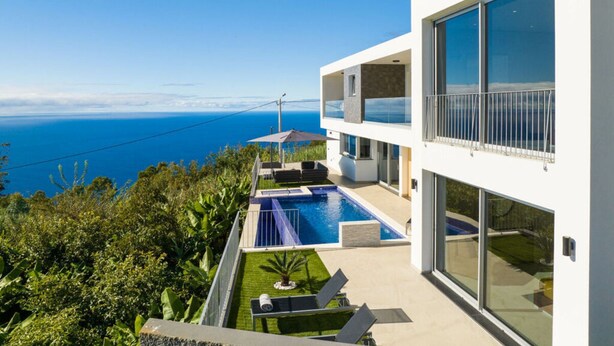 A modern two-story building with a rooftop pool, large windows, and a view of the ocean.