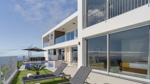 A modern two-story building with a balcony, a swimming pool, and two lounge chairs.
