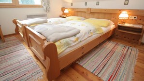A cozy bedroom with a wooden bed, two bedside lamps, a window with floral curtains, and a small wooden nightstand. - Apartment in Tropolach With Sauna (Hermagor-Pressegger See)