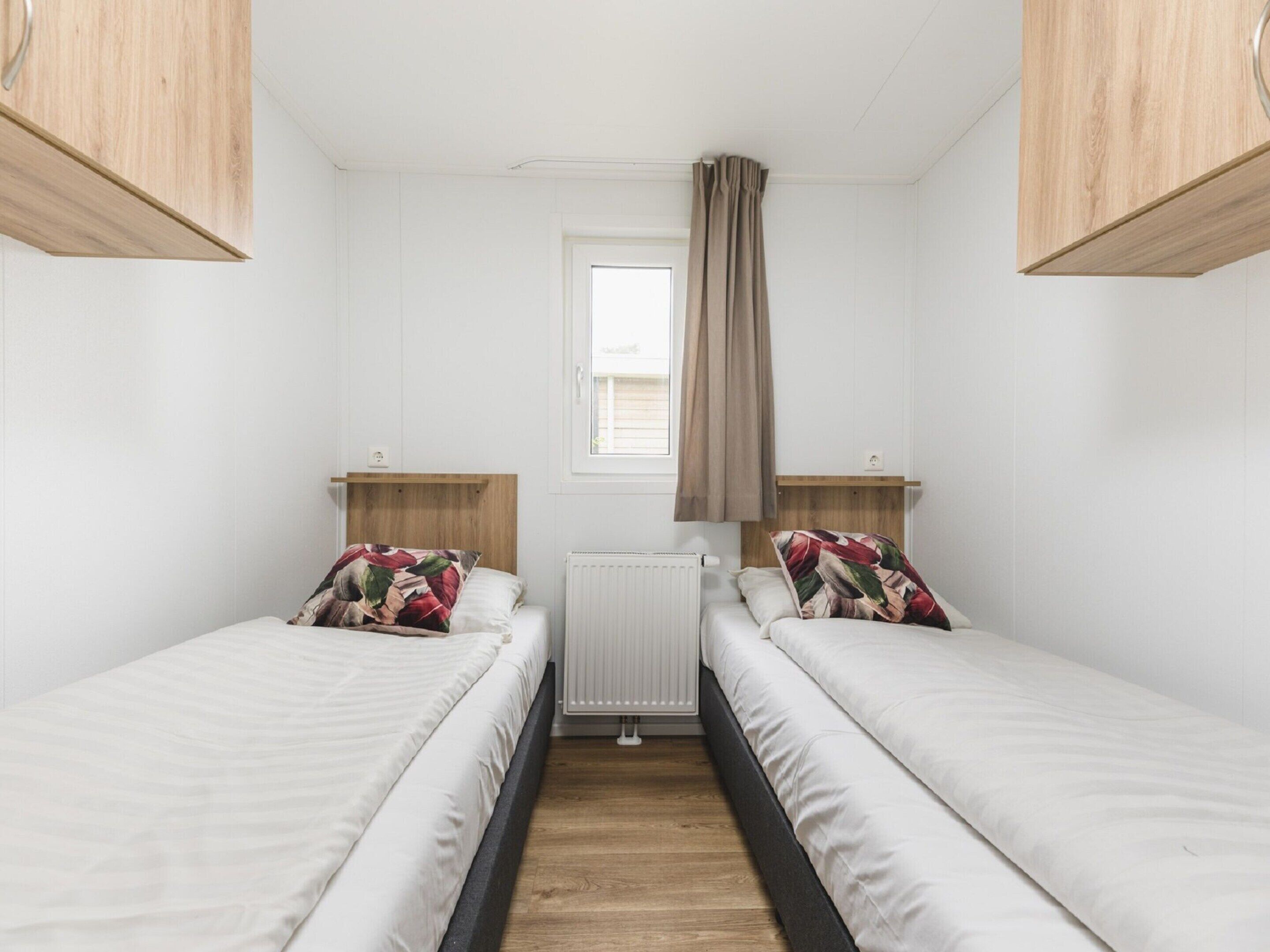 A small, clean room with two single beds, wooden headboards, a window with a curtain, and a radiator.
