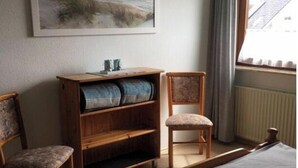 A bedroom with a bed, a wooden nightstand, two chairs, a bookshelf, and a painting of a beach scene. - Holiday Home Next Door to Busum (Büsum)