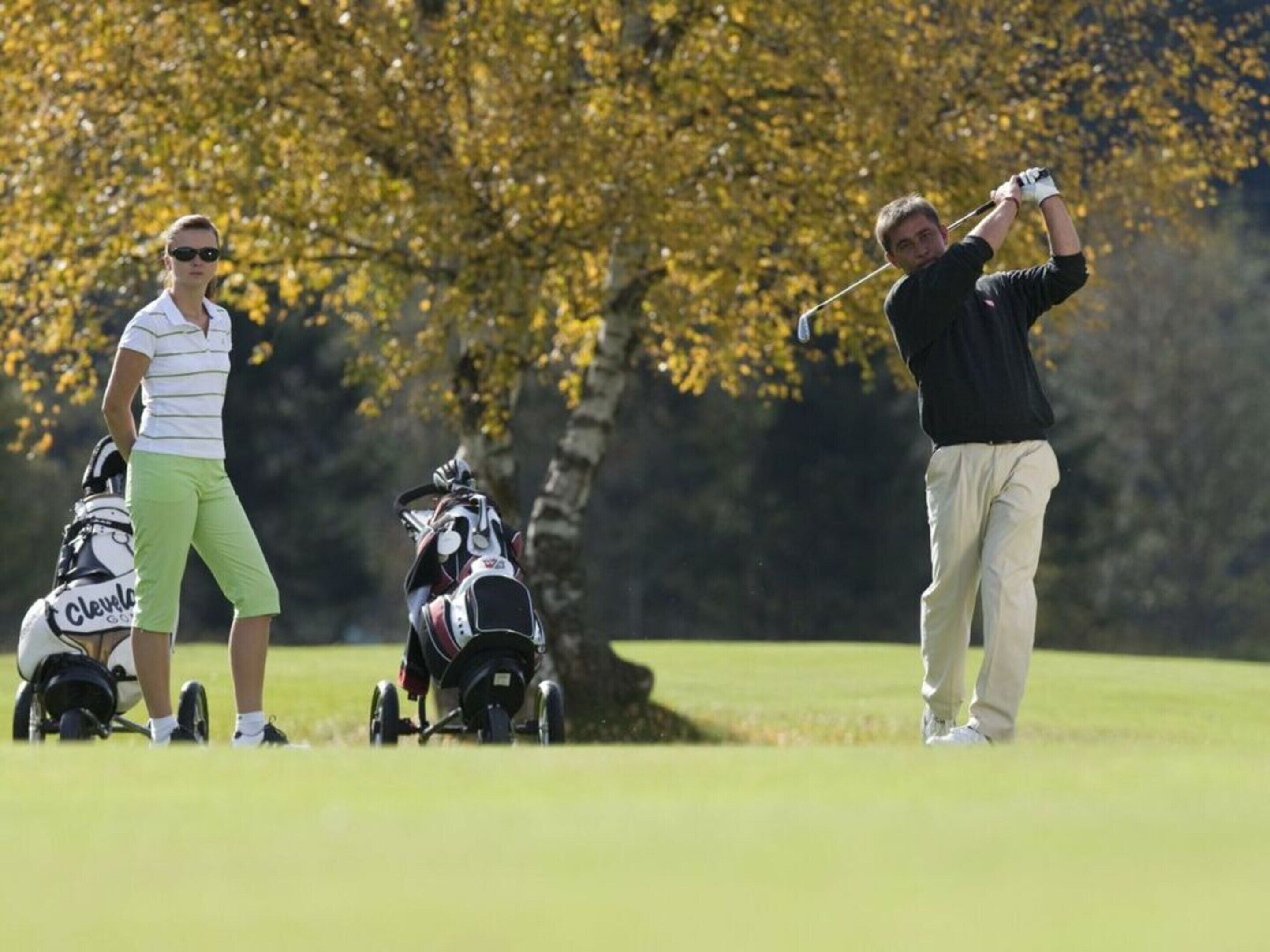 Un terrain de golf avec un joueur en plein élan, un golfeur avec un sac de clubs et un autre golfeur avec un sac de clubs.