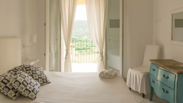 Quarto com cama, uma cadeira, cômoda e vista para uma varanda.