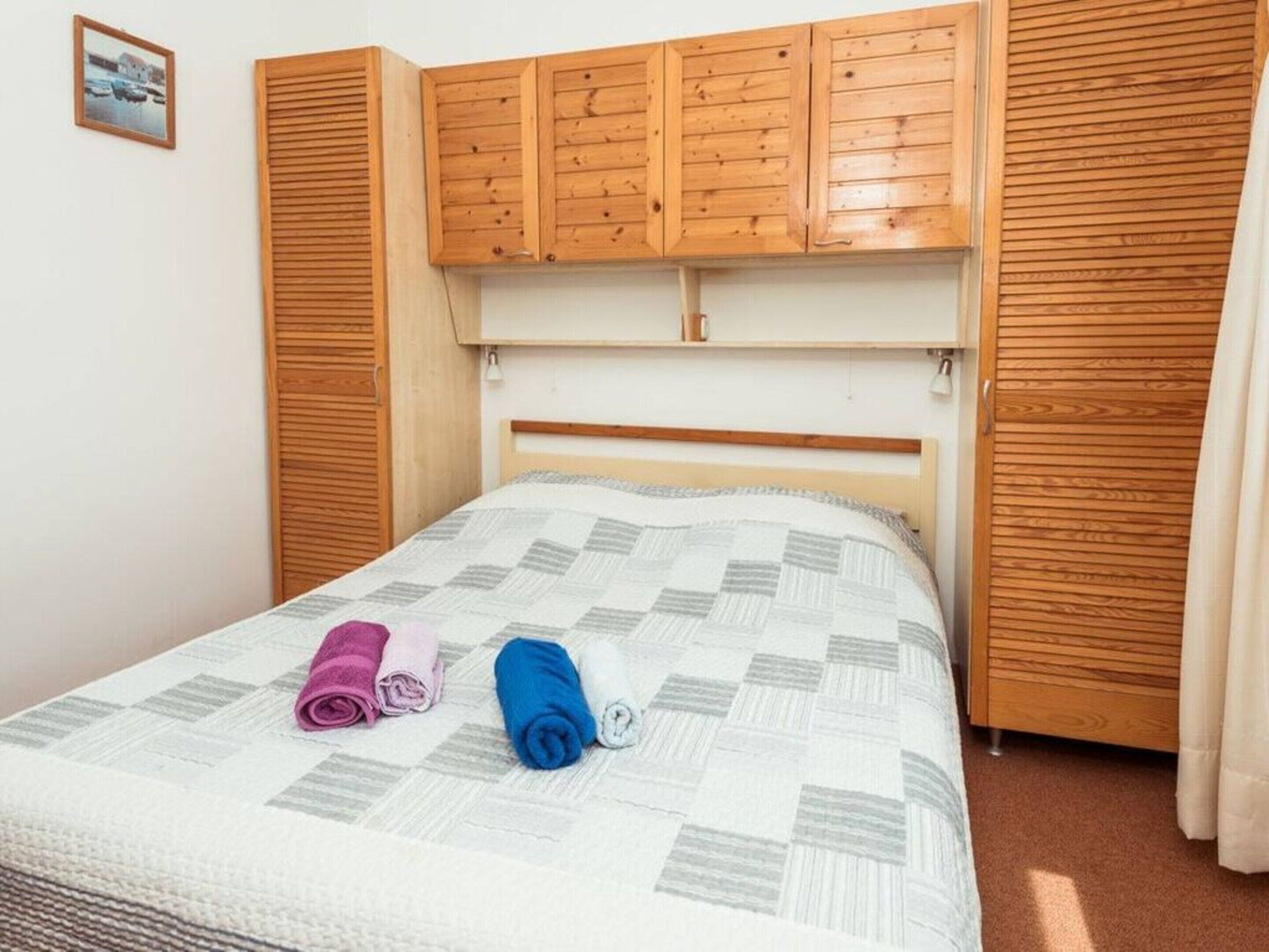 Egy gondosan megterített ágy, rajta egy takaró, fából készült ágyvég és egy tükörrel ellátott, szintén fából készült szekrény.