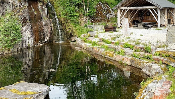 A rustic wooden cabin with a stone pathway leading to it, a small waterfall, and a calm pond. - 8 Person Holiday Home in Valldal (Fjord)