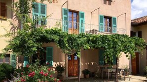 A building with a covered patio area, a gravel ground, and potted plants with pink flowers. - Holiday Home Casa Nanda (Mombaruzzo)