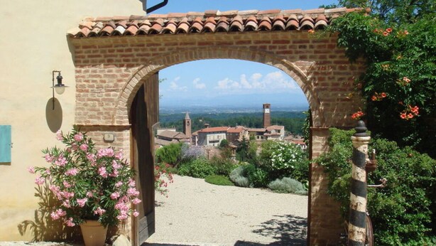 An arched stone gateway leading to a cobblestone path, surrounded by lush greenery and a view of a distant town with a prominent clock tower. - Holiday Home Casa Nanda (Mombaruzzo)