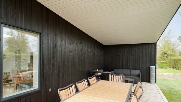 A modern outdoor dining area with a wooden table and metal chairs, a grill, and a view of the garden. - 5 Star Holiday Home in Nysted (Nysted)