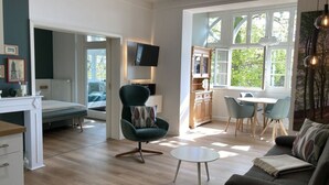 Una sala de estar moderna con un sofá, una mesa redonda de centro y un sillón verde con un cojín estampado. Hay una zona de comedor con mesa y sillas, y una cocina con armarios de madera y horno empotrado. Un televisor de pantalla plana está montado sobre el sofá, y una ventana proporciona luz natural.