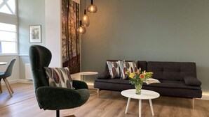 Una sala moderna con un sofá oscuro, un sillón verde, una mesita blanca y un jarrón con flores.
