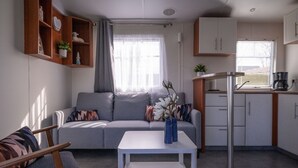 Moderni olohuone, jossa on harmaa sohva, valkoinen sohvapöytä, jossa on lehtiä, valkoisilla kaapeilla varustettu keittiöalue ja kahvinkeitin sekä ikkuna, jossa on sälekaihtimet.