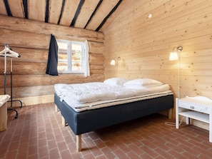 A bedroom with a bed, a nightstand, a lamp, a window with curtains, and a wooden wall. - 24 Person Holiday Home in Laeso-by Traum (Læsø)