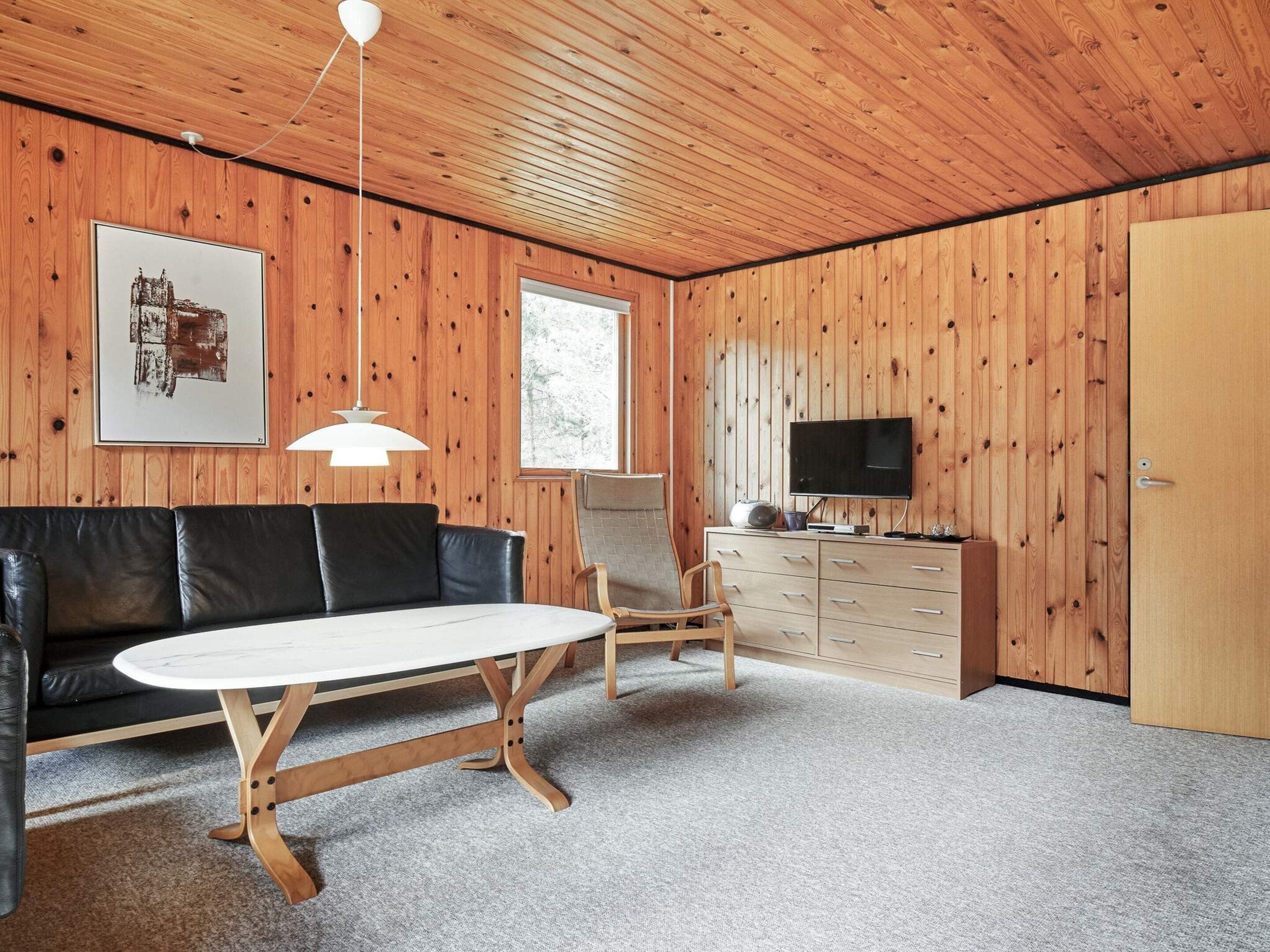 Une pièce avec un plafond en bois, un canapé en cuir noir, une table ronde, une chaise en bois, un téléviseur posé sur un meuble en bois et un tableau encadré accroché au mur.