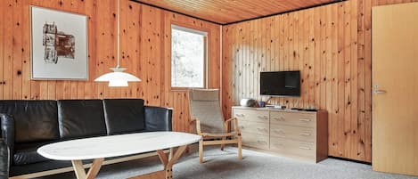 Una habitación con techo de madera, un sofá de cuero negro, una mesa redonda, una silla de madera, un televisor sobre un mueble de madera y un cuadro enmarcado en la pared.