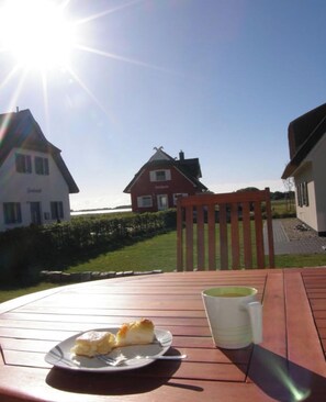 Ein Holztisch mit Essen auf einem Teller und einer Tasse, ein Holzstuhl sowie im Hintergrund Häuser und ein Hecke.