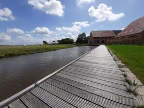 Un paseo marítimo de madera al lado de un canal, con ovejas pastando al fondo y un edificio de techo rojo.