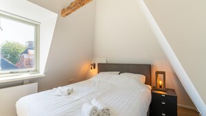 A bedroom with a sloped ceiling, a bed with white linens, a bedside table with a lamp, and a view of buildings outside the window. - 2 Adjacent Apartments in the Heart of Sneek (Sneek)